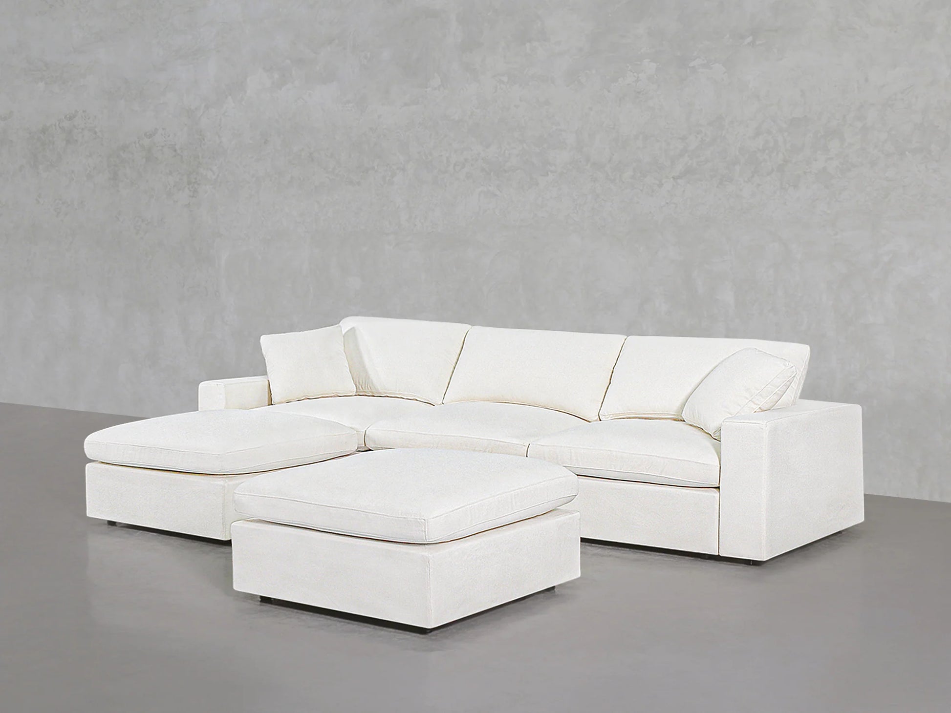 A modern 4-seat modular chaise sectional with an ottoman is set against a minimalist gray wall and floor, featuring clean lines and plush cushions.