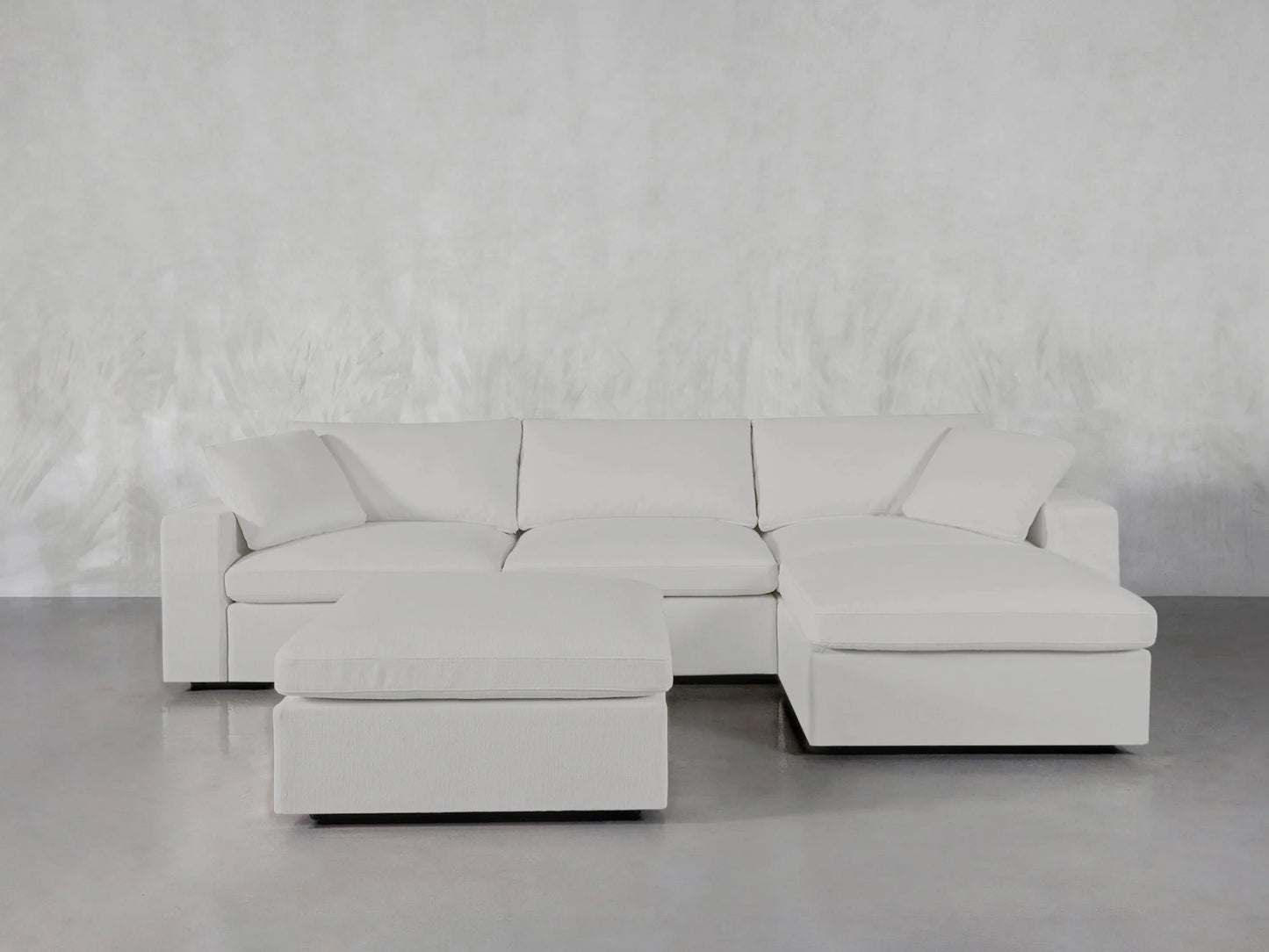 In a minimalist room with a light gray floor and an abstract background wall, the 4-Seat Modular Chaise Sectional with Ottoman is elegantly displayed. This modern white sectional showcases clean lines, providing a generous seating area.