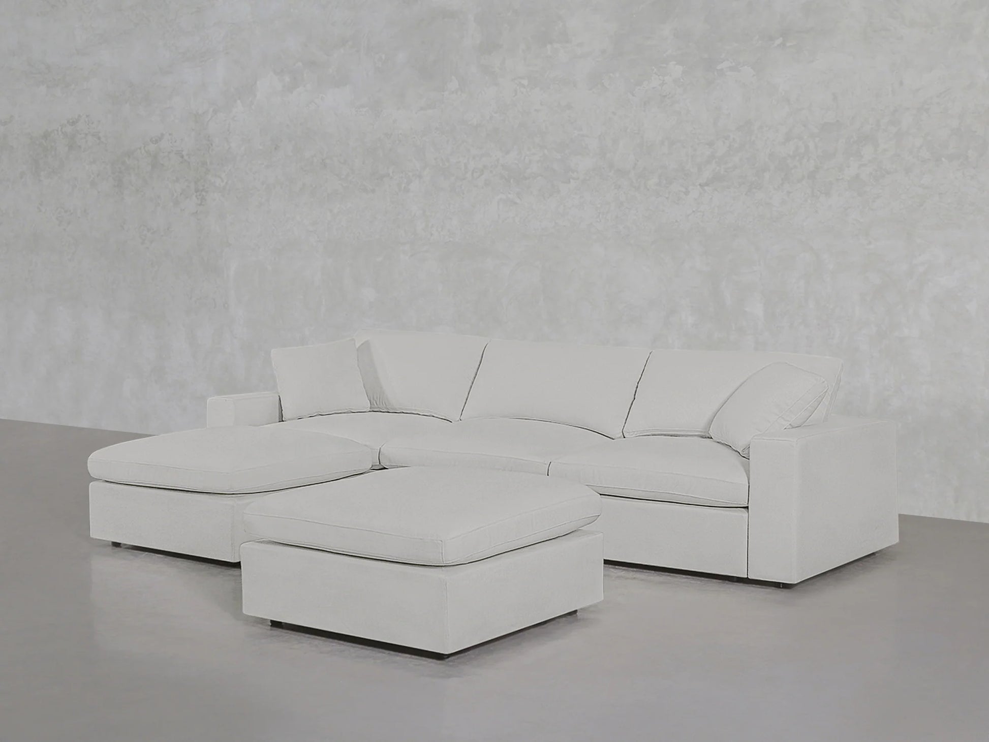 A 4-Seat Modular Chaise Sectional with Ottoman in a minimalist white design is set on a sleek concrete floor, contrasted by a textured light gray wall.