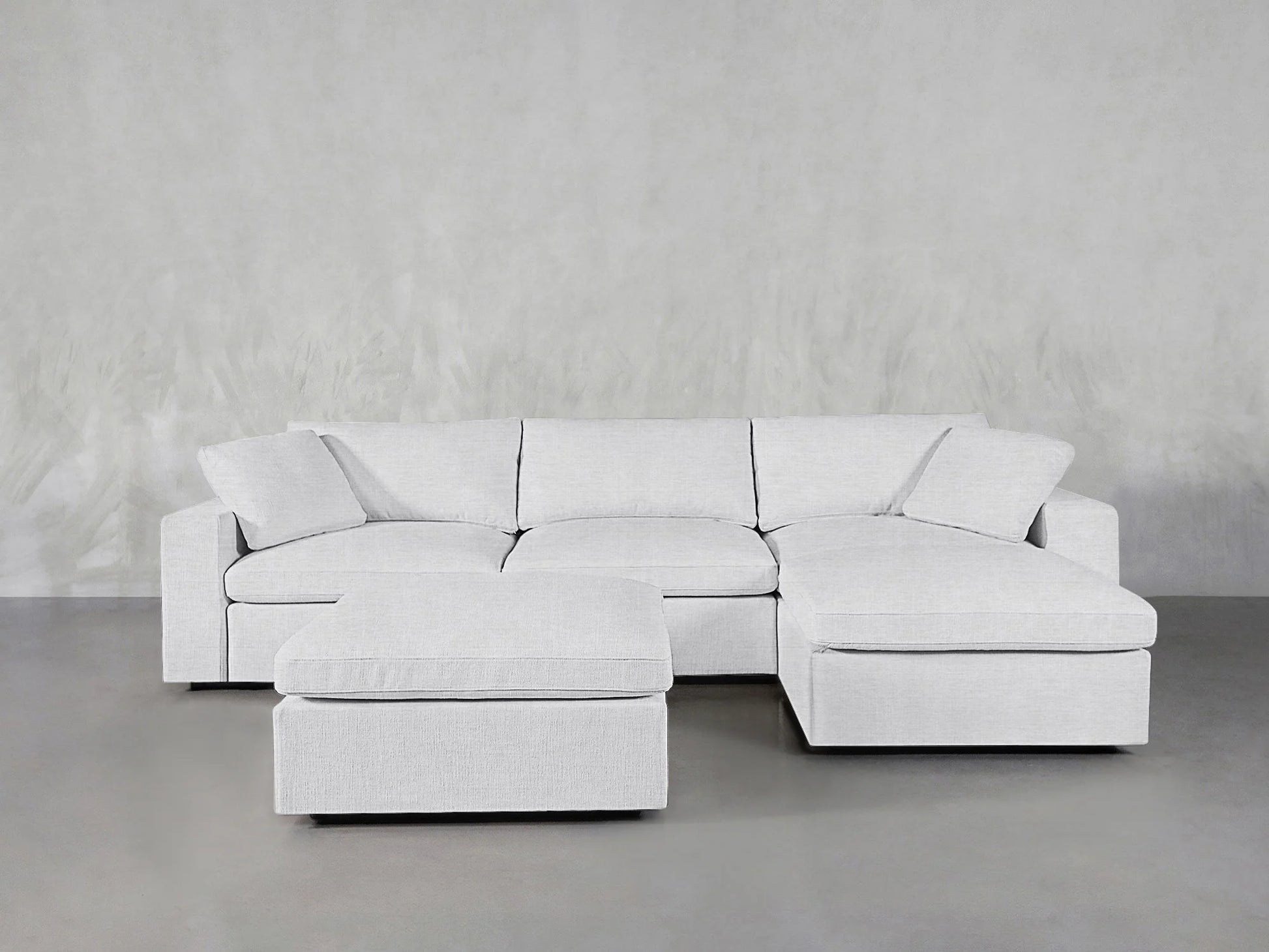 A contemporary, light gray 4-seat modular chaise sectional with an ottoman is elegantly arranged on a smooth gray floor, complemented by a textured gray wall. The sectional boasts plush cushions for added comfort.