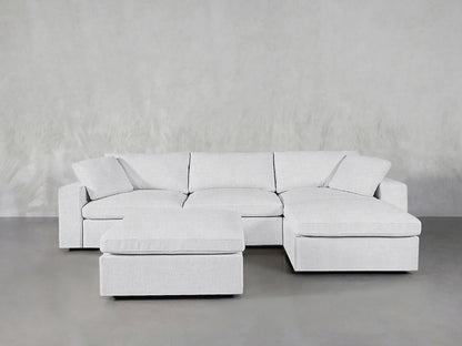 A contemporary, light gray 4-seat modular chaise sectional with an ottoman is elegantly arranged on a smooth gray floor, complemented by a textured gray wall. The sectional boasts plush cushions for added comfort.