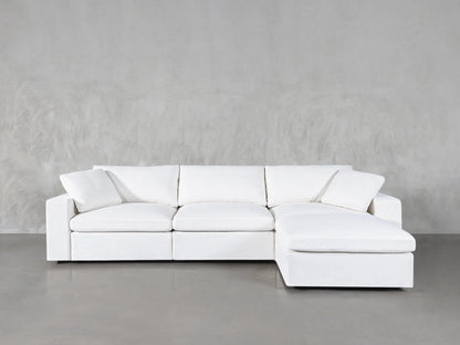 The 4-Seat Modular Chaise Sectional, with its modern design featuring clean lines and plush cushions, is elegantly positioned against a light gray wall on a smooth concrete floor. This white sectional includes a chaise on the right side, offering both spaciousness and minimalist comfort.