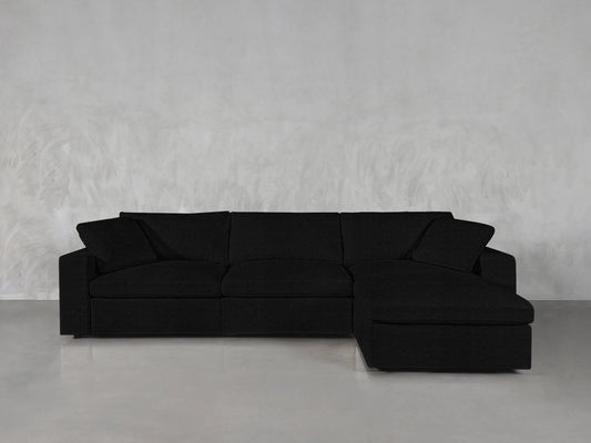 A modern 4-Seat Modular Chaise Sectional in black is placed against a plain light gray wall. The sectional boasts clean lines and a minimalist design, resting on a smooth, light-colored floor.