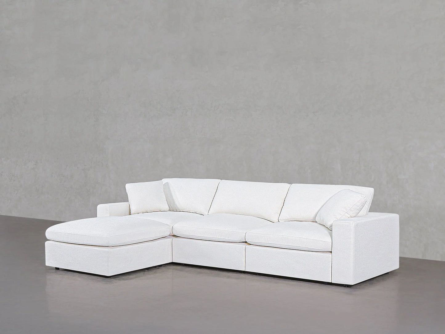 A modern 4-Seat Modular Chaise Sectional in white is placed against a plain gray wall on a polished concrete floor. This sectional features thick cushions and clean lines, achieving a minimalist appearance.