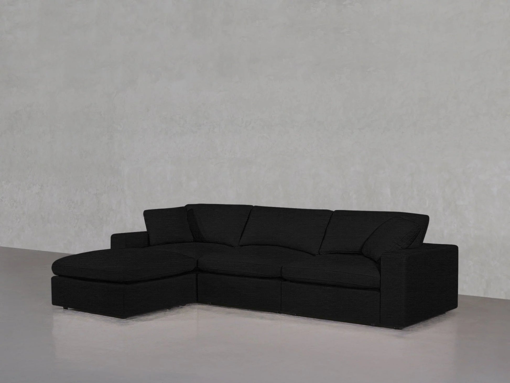 A contemporary, dark gray 4-Seat Modular Chaise Sectional with plush cushions is arranged against a minimalist light gray concrete wall, featuring an L-shaped configuration with a chaise lounge on the left-hand side.