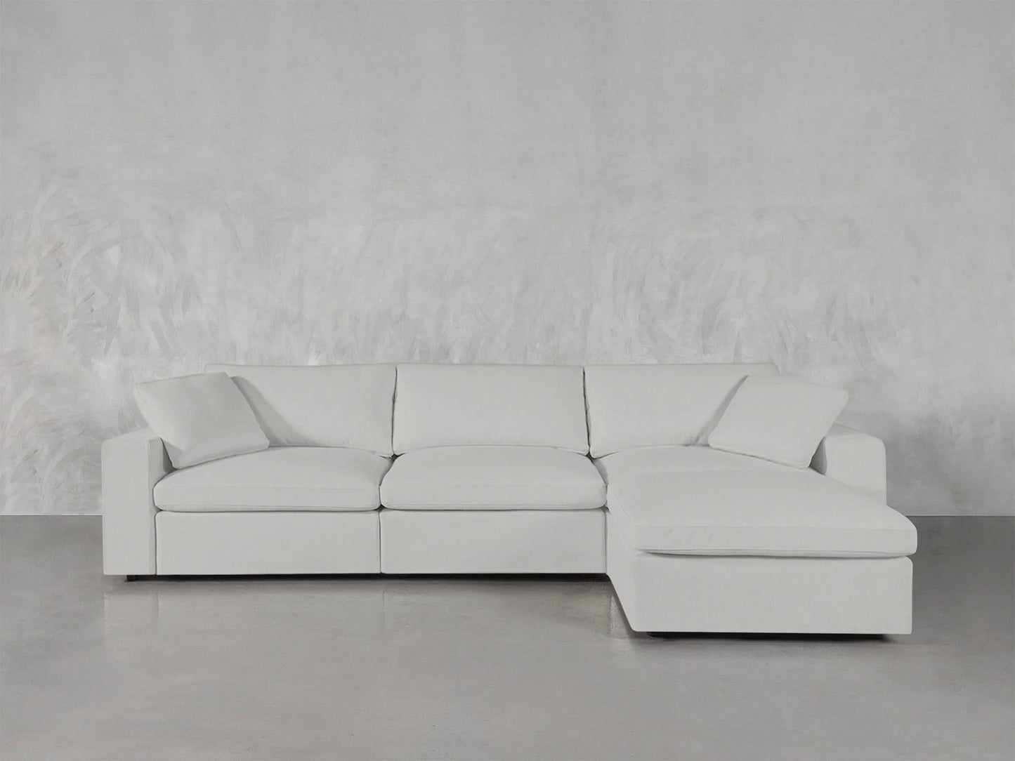 A minimalist 4-Seat Modular Chaise Sectional in light gray is set against a matching textured wall, with the chaise positioned on the right. It's placed on a smooth, concrete floor, with two cushions adding to its modern aesthetic.