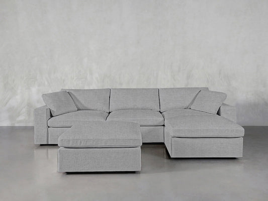 A contemporary 4-seat modular chaise sectional with ottoman is placed against a minimalist light gray wall on a polished concrete floor. The sectional includes a chaise lounge on the right side, enhancing its modern and sleek design.