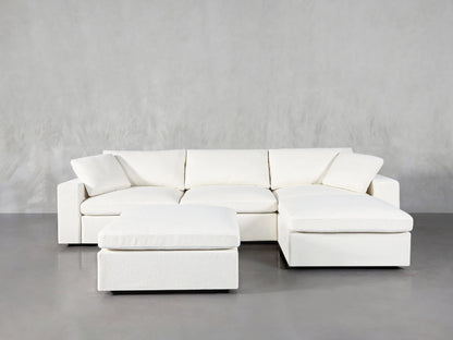 A contemporary, minimalist living room showcasing a 4-Seat Modular Chaise Sectional with Ottoman in white, featuring a chaise on the right. The space maintains a light, neutral color palette with sleek gray flooring and an unadorned gray wall as the backdrop.