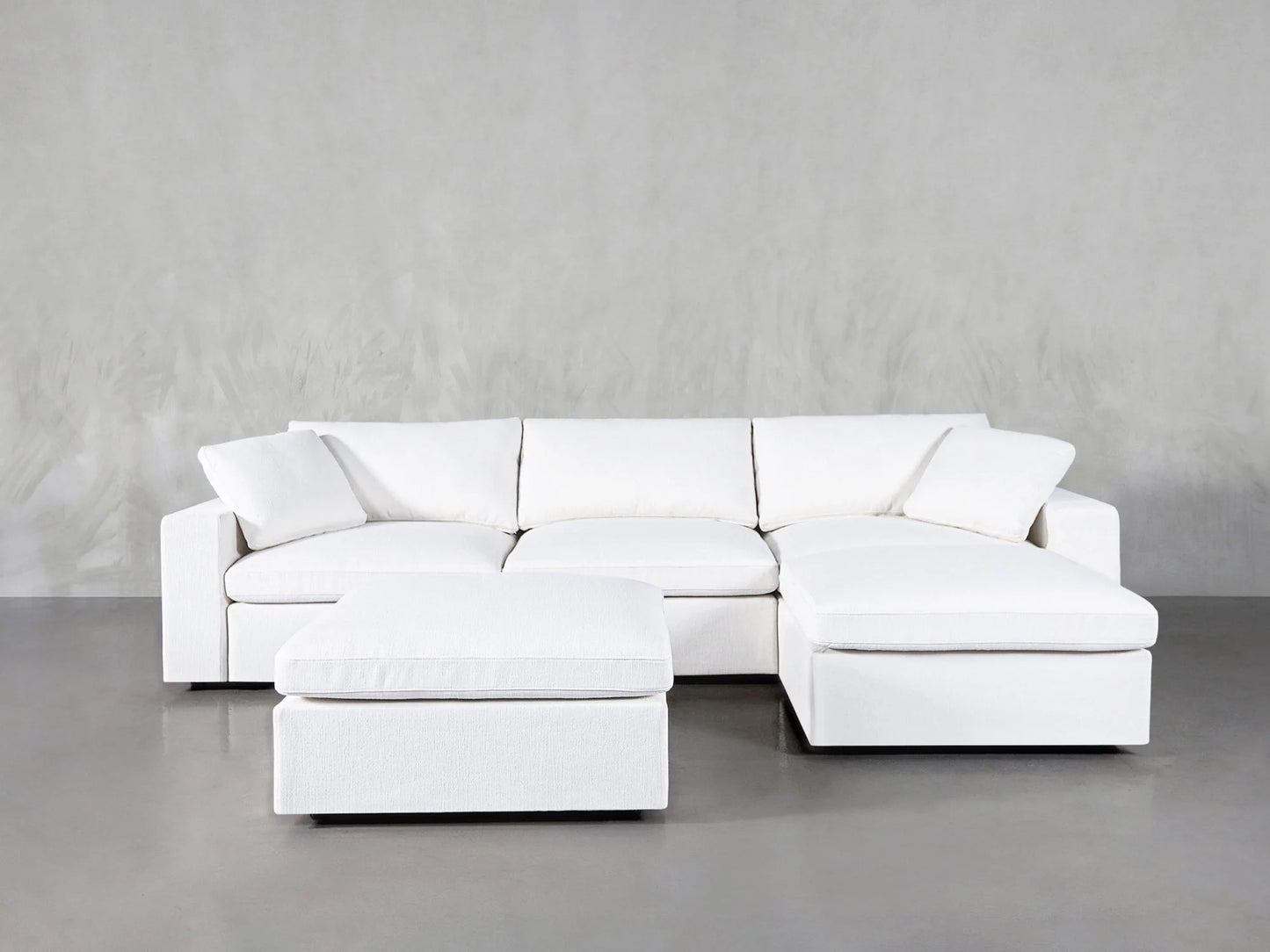 The 4-Seat Modular Chaise Sectional with Ottoman, featuring plush cushions and a modern white design, is displayed against a plain gray background. The coordinating ottoman is placed in front of one section, enhancing the stylish and minimalist setup.