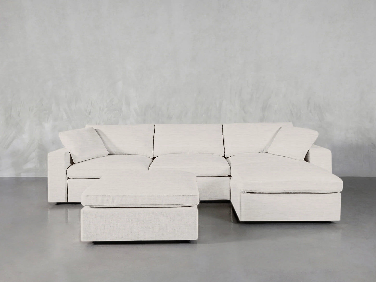 The 4-Seat Modular Chaise Sectional with Ottoman in cream features a chaise lounge on the right side. It is set against a smooth gray floor, complemented by a subtly patterned gray wall in the background.