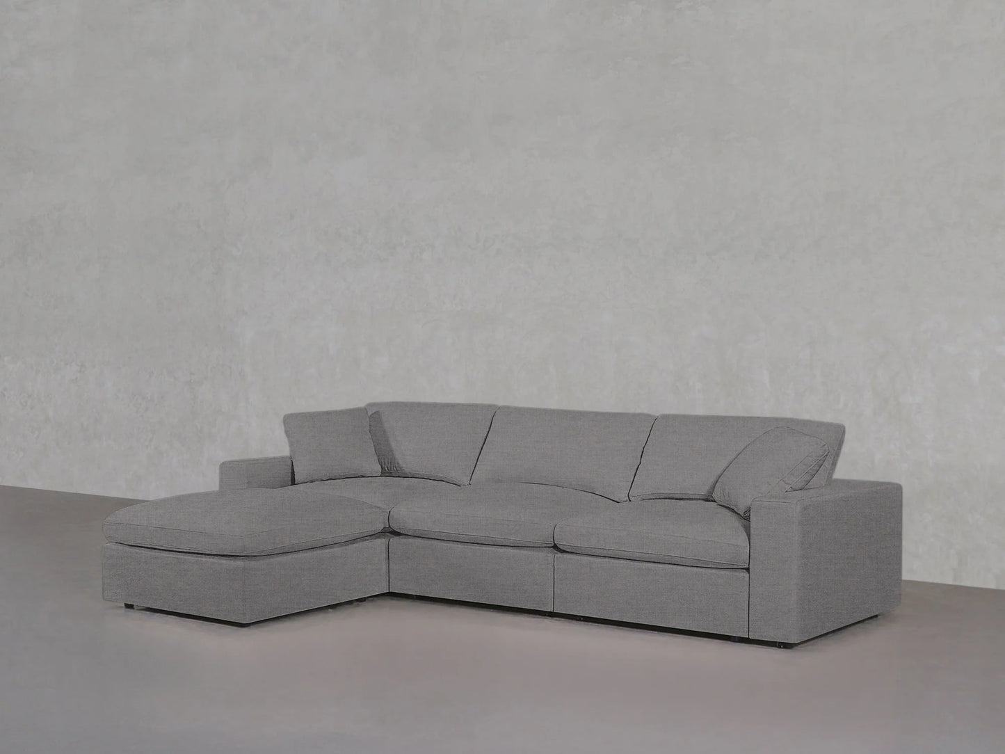 A 4-Seat Modular Chaise Sectional in a modern gray hue, featuring a left-side chaise, complements the plain light gray textured wall. With its clean lines and plush cushions, the sectional presents a minimalist and comfortable look.