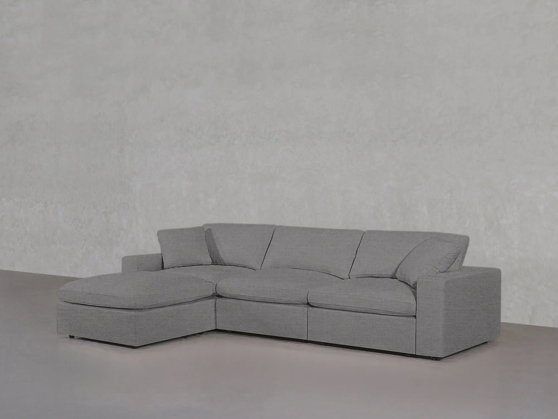 A 4-Seat Modular Chaise Sectional in a modern gray hue, featuring a left-side chaise, complements the plain light gray textured wall. With its clean lines and plush cushions, the sectional presents a minimalist and comfortable look.