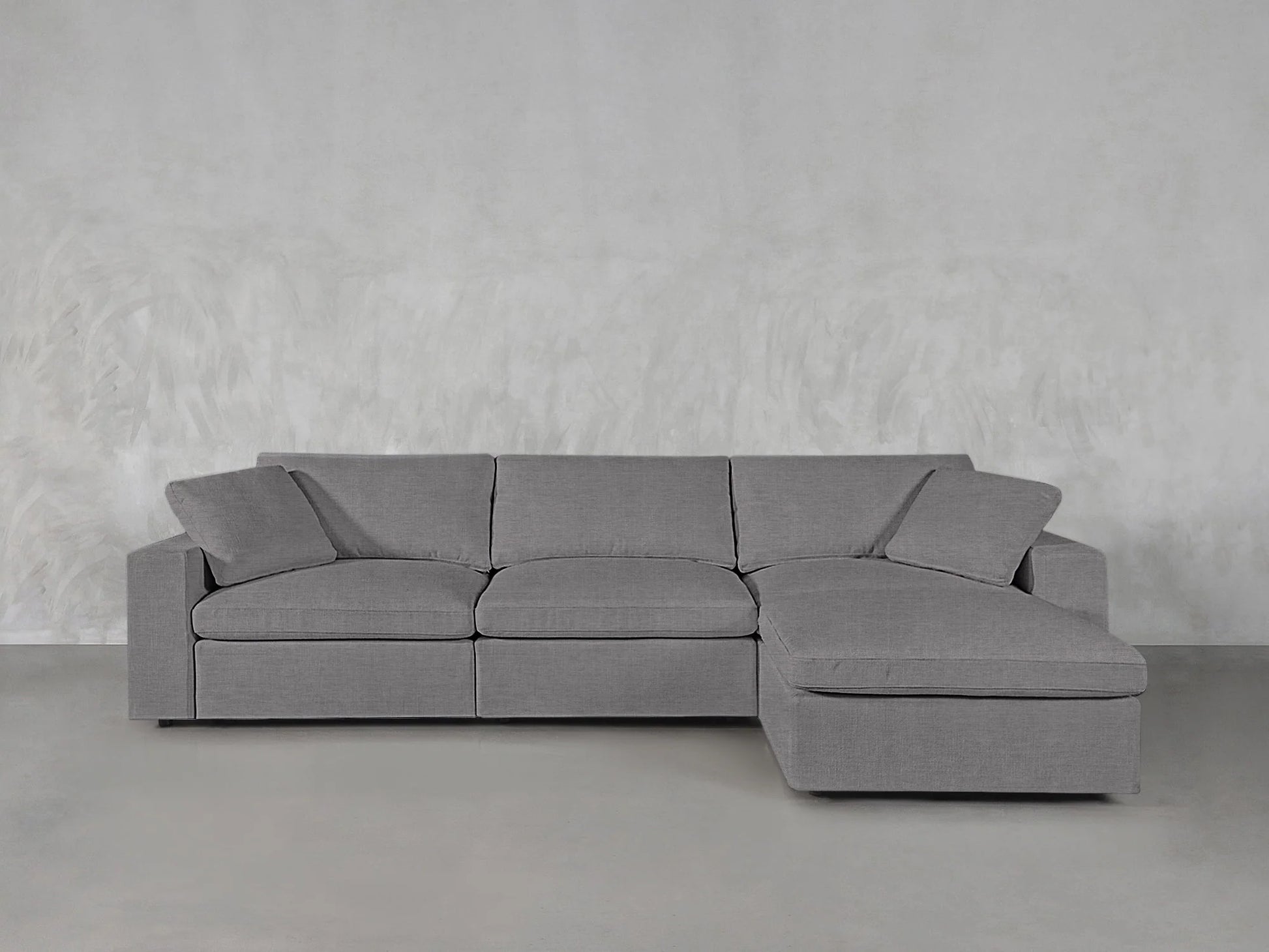 The 4-Seat Modular Chaise Sectional, in gray, features a right-facing chaise lounge positioned against a light textured backdrop. It includes three seat cushions and matching backrest cushions to create a minimalist and modern appearance.