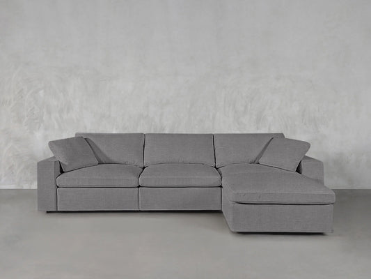 The 4-Seat Modular Chaise Sectional, in gray, features a right-facing chaise lounge positioned against a light textured backdrop. It includes three seat cushions and matching backrest cushions to create a minimalist and modern appearance.