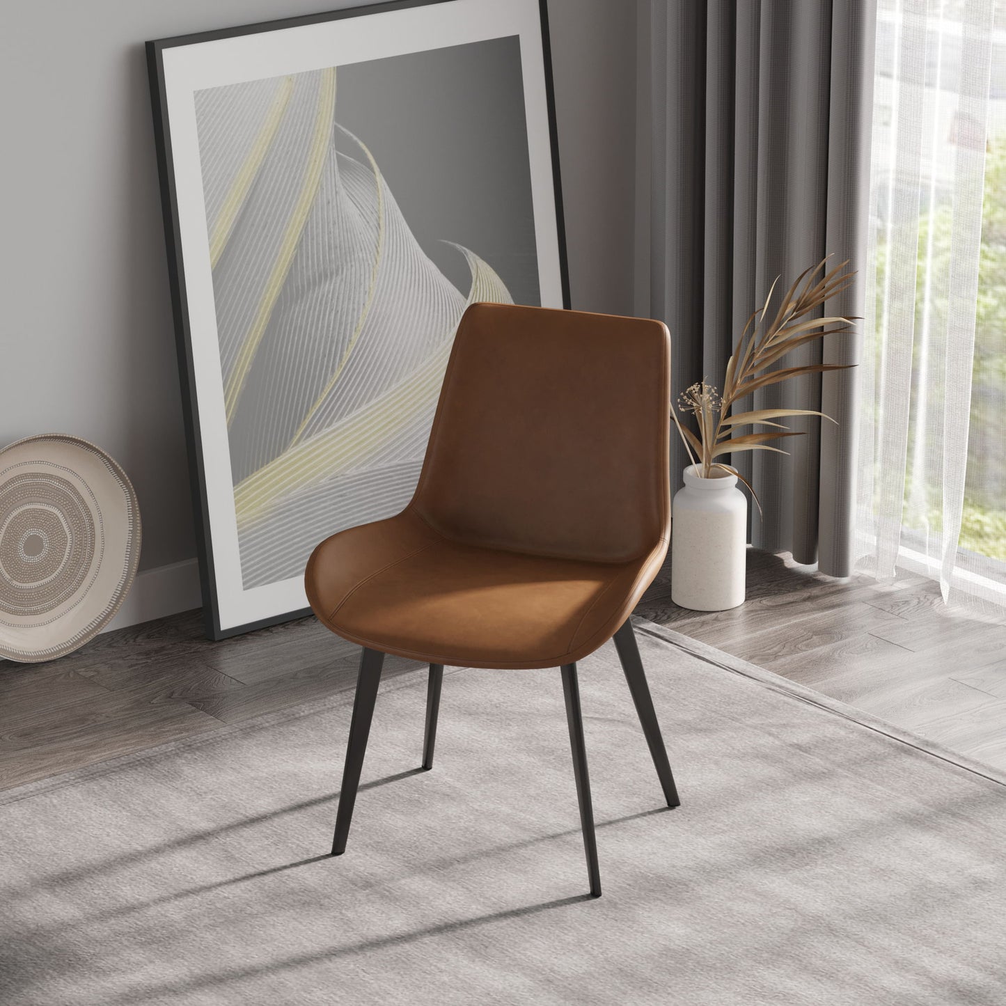 A modern room includes a Modern Dining Chair with brown leather and black metal legs on a gray carpet. A framed abstract artwork leans against the wall, while a vase of dried plants is nearby. Sheer curtains adorn the window in the background.