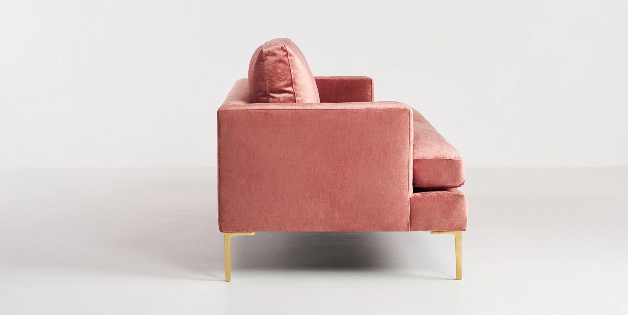 Side view of the Bowen 91" Sofa, a modern pink velvet piece with gold metal legs, set against a white background. The sofa boasts a minimalist design with its boxy shape and plush cushions.