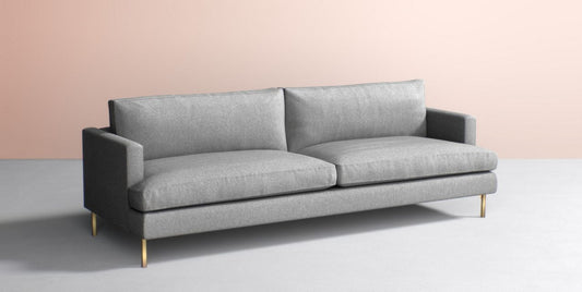 The Bowen 91" Sofa, with its modern design, features a sleek grey finish and clean lines. It includes two large cushions and is complemented by slim, angular armrests and elegant gold-colored legs. Placed against a soft pink wall and light grey floor, it adds a sophisticated touch to the room.