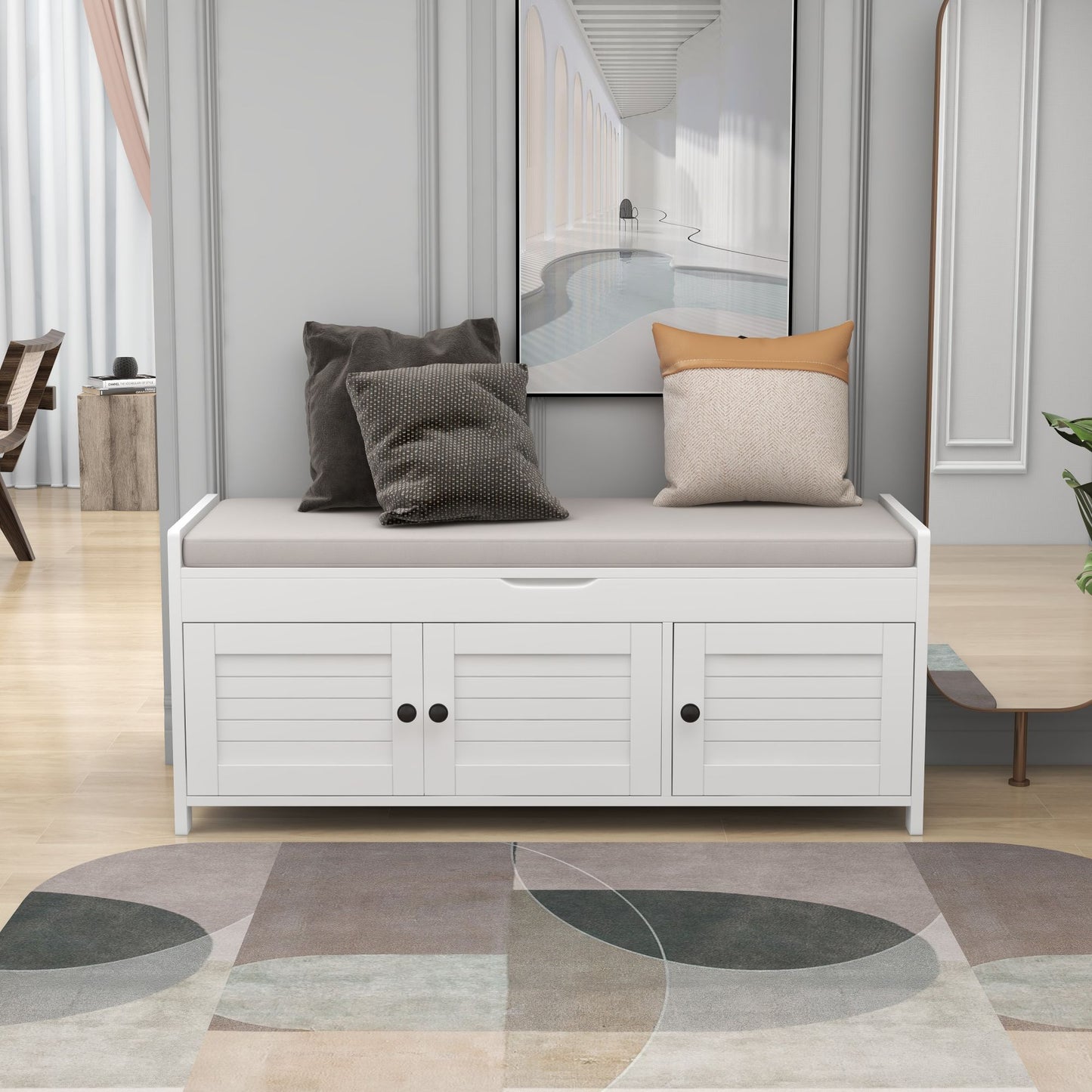 A stylish entryway is highlighted by a storage bench with three shutter-shaped doors, a removable cushioned seat, and hidden storage. On top rest two gray and beige pillows. A neutral-toned rug and modern wall art enhance the contemporary decor.