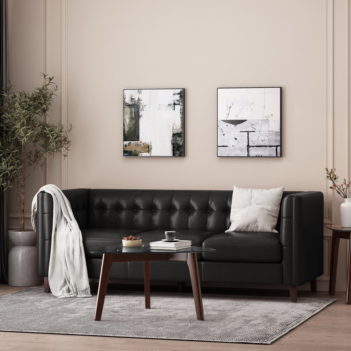 A modern living room features a black Comfy 3 Seat Sofa with tufted back, a white throw pillow, and blanket draped over the arm. A wooden coffee table holds a cup, jug, and plate. Abstract paintings adorn the beige wall, and a potted plant sits in the corner.