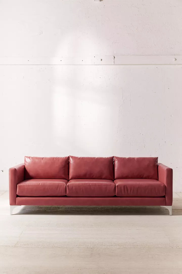 The Chamberlin Recycled Leather Sofa, a red, three-seater with plush cushions, is positioned against a white, textured wall. Sunlight casts a gentle glow on parts of the wall, creating a warm and inviting atmosphere. The floor is light-colored wood.