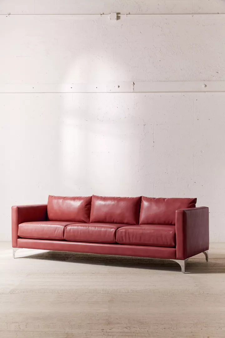 A Chamberlin Recycled Leather Sofa with three cushions is elegantly placed in a minimalistic room, where the background showcases a white textured wall and light wood flooring.