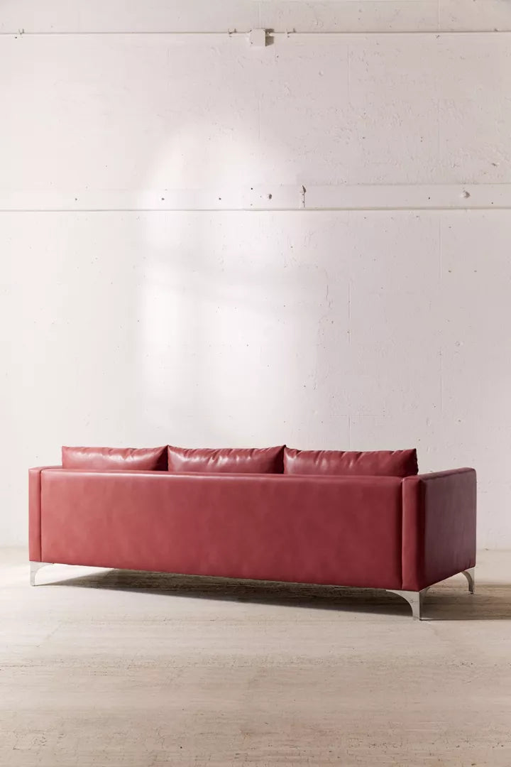 The Chamberlin Recycled Leather Sofa, featuring a modern design in red with three back cushions, sits against a white, textured wall. Its sleek metal legs elevate the sofa on a light wood floor, enhancing the minimalist aesthetic.