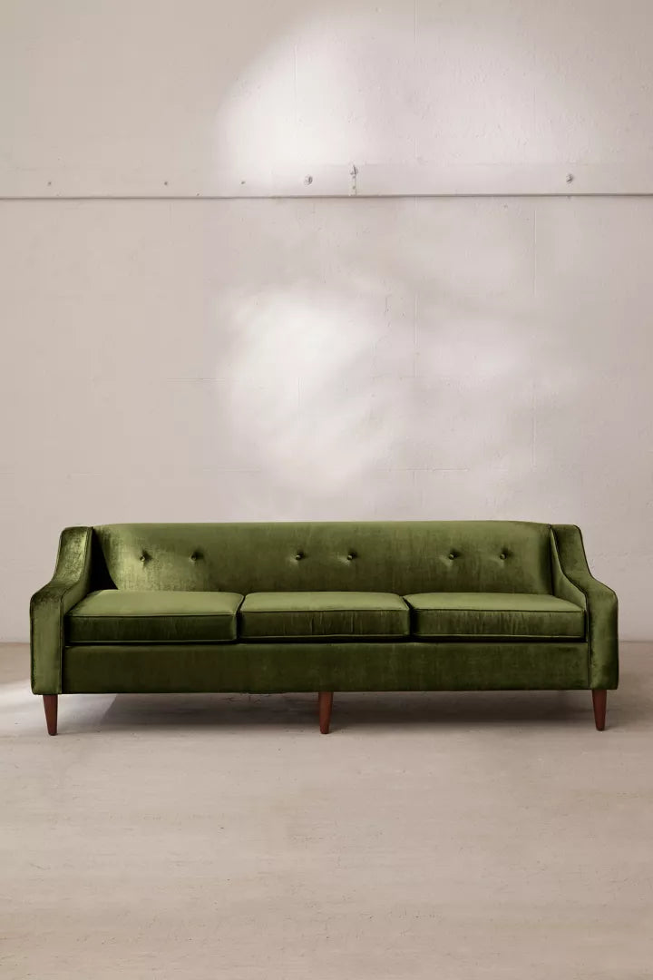 A Milly Velvet Sofa in green is placed against a plain wall, featuring three cushions with wooden legs and button detailing on the back cushions. Soft lighting casts subtle shadows on the wall behind it.