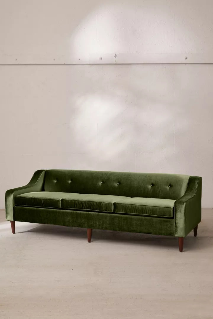 The Milly Velvet Sofa, in a vibrant green velvet fabric with a mid-century modern design, stands against a neutral wall. It showcases button-tufted back cushions and elegant wooden legs. Sunlight casts soft shadows on the wall and floor.