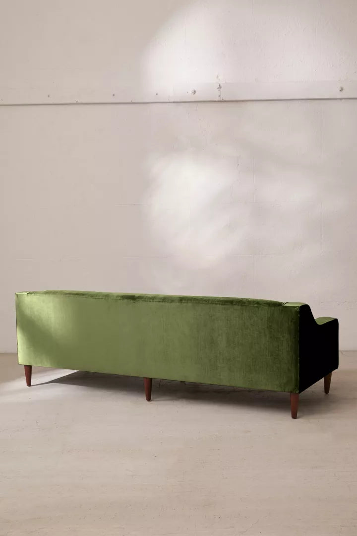 The Milly Velvet Sofa in green, featuring wooden legs, is placed against a light-colored wall. Its sleek, modern design showcases clean lines and a minimalist aesthetic. Natural light casts subtle reflections on the wall.