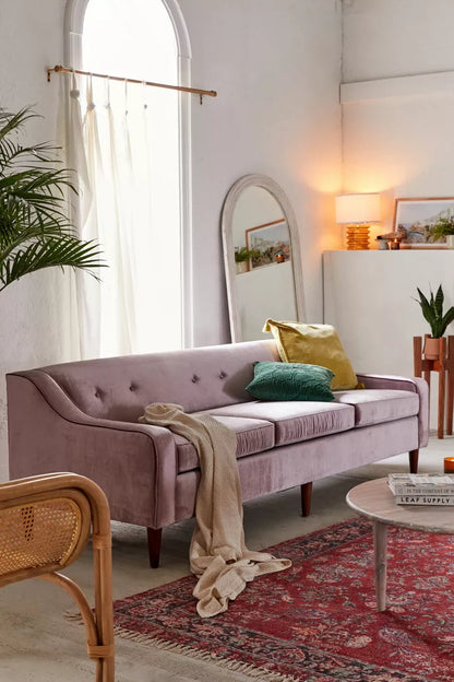 A cozy living room featuring the Milly Velvet Sofa in lavender, elegantly decorated with green and gold pillows, a draped blanket, and complemented by a round wooden coffee table. A tall mirror, a potted plant, and warm lighting enhance the inviting atmosphere. The floor is covered by a red patterned rug.