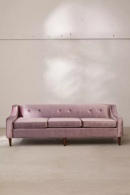 The Milly Velvet Sofa, a three-seater in pink velvet with tufted cushions and wooden legs, is positioned against a white wall. Natural light casts soft shadows on the wall behind the sofa.