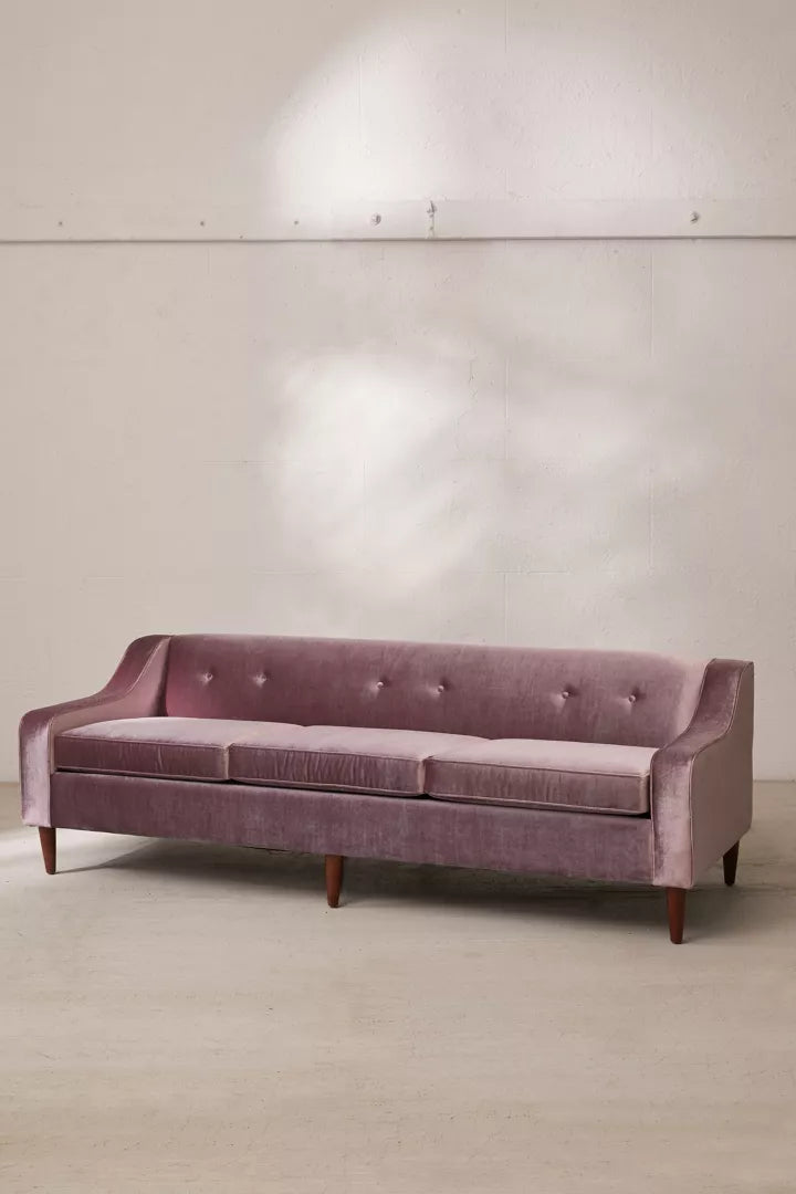 The Milly Velvet Sofa in a muted pink velvet finish, featuring wooden legs, is positioned against a plain, light gray wall. This three-seater couch includes a tufted backrest, smooth cushions, and angular armrests that cast soft shadows on the floor.