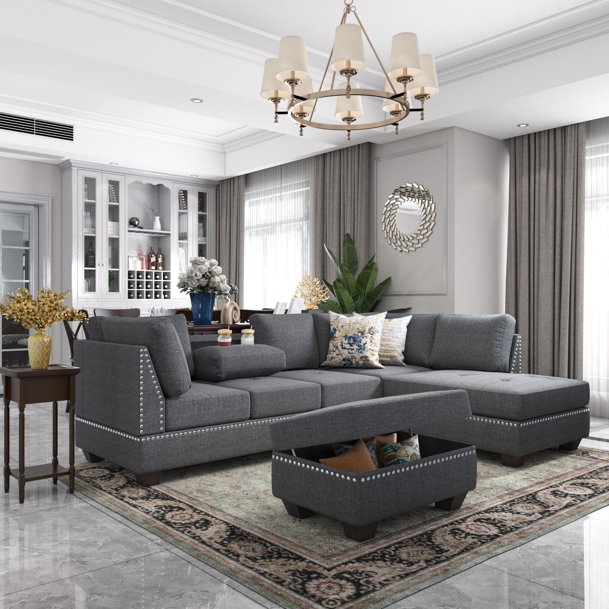 A modern living room features a Reversible Sectional Sofa Space Saving with Storage Ottoman. It includes patterned cushions and a chandelier, beige curtains on large windows, a decorative mirror, plants, and a wooden side table adorned with flowers.