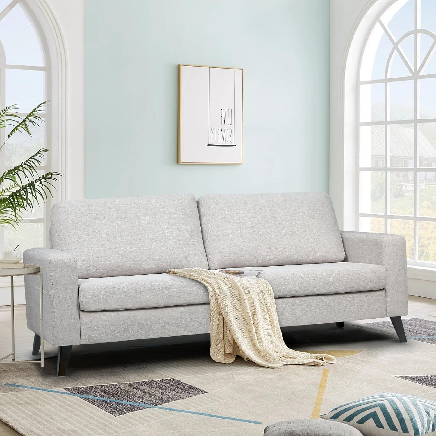 A modern living room features the YODOLLA 79" Sectional Sofa in a light gray shade with a cream blanket draped over it. The room is complemented by pastel blue walls, large arched windows, a geometric rug, and a potted plant. A minimalistic framed artwork adorns the wall.