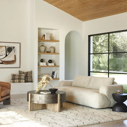 A modern living room features the Benito Sofa by Four Hands in a beige hue, accompanied by a round coffee table and a brown armchair. A large window allows natural light to pour in. Built-in shelves display decorative items, while abstract art adorns the walls, and a textured rug lays across the wooden floor.