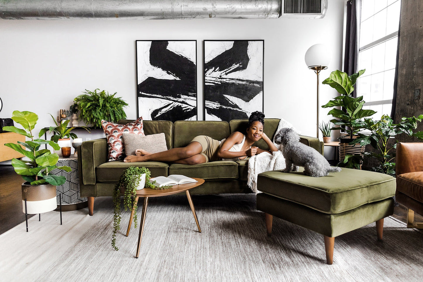 Someone relaxes on an Albany Sofa in Olive Velvet, smiling at a small dog in a contemporary living room. The space features large plants, abstract black-and-white artwork, and a round wooden coffee table with a book atop it. Sunlight pours through a large window.