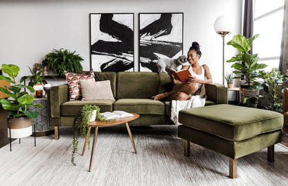 A woman sits on an Albany Sofa in olive velvet, reading a book in a modern living room adorned with plants and abstract artwork on the wall. A small dog is beside her. In the foreground, there is a matching ottoman and a round wooden coffee table topped with books.