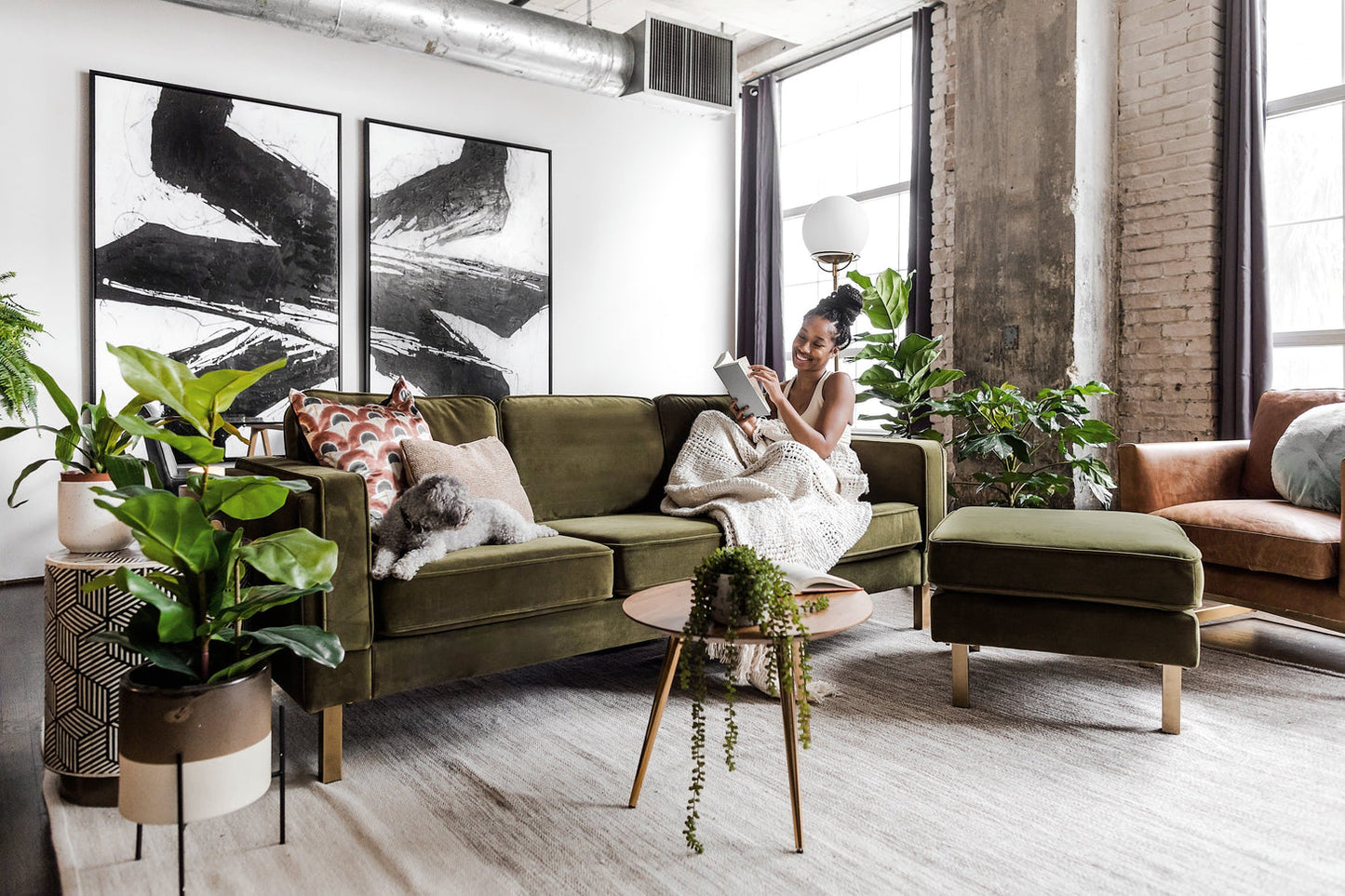 A woman sits on an Albany Sofa in olive velvet in a bright, modern living room, reading a book. A dog lies beside her. The room features large abstract artwork, houseplants, and a mix of industrial and cozy decor elements.