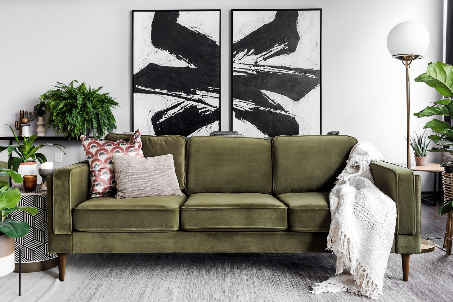 An updated living room features an Albany Sofa in Olive Velvet, complemented by abstract black-and-white wall art and assorted plants. A textured throw blanket elegantly drapes over the sofa, while a geometric side table displays decorative items. Nearby, a floor lamp stands on wooden flooring.