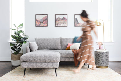 A woman in a brown dress walks past a stylish living room featuring the Park Sofa in grey fabric, adorned with colorful pillows and illuminated by a modern floor lamp. Three paintings hang on the white wall, and a large plant is positioned beside the sofa.