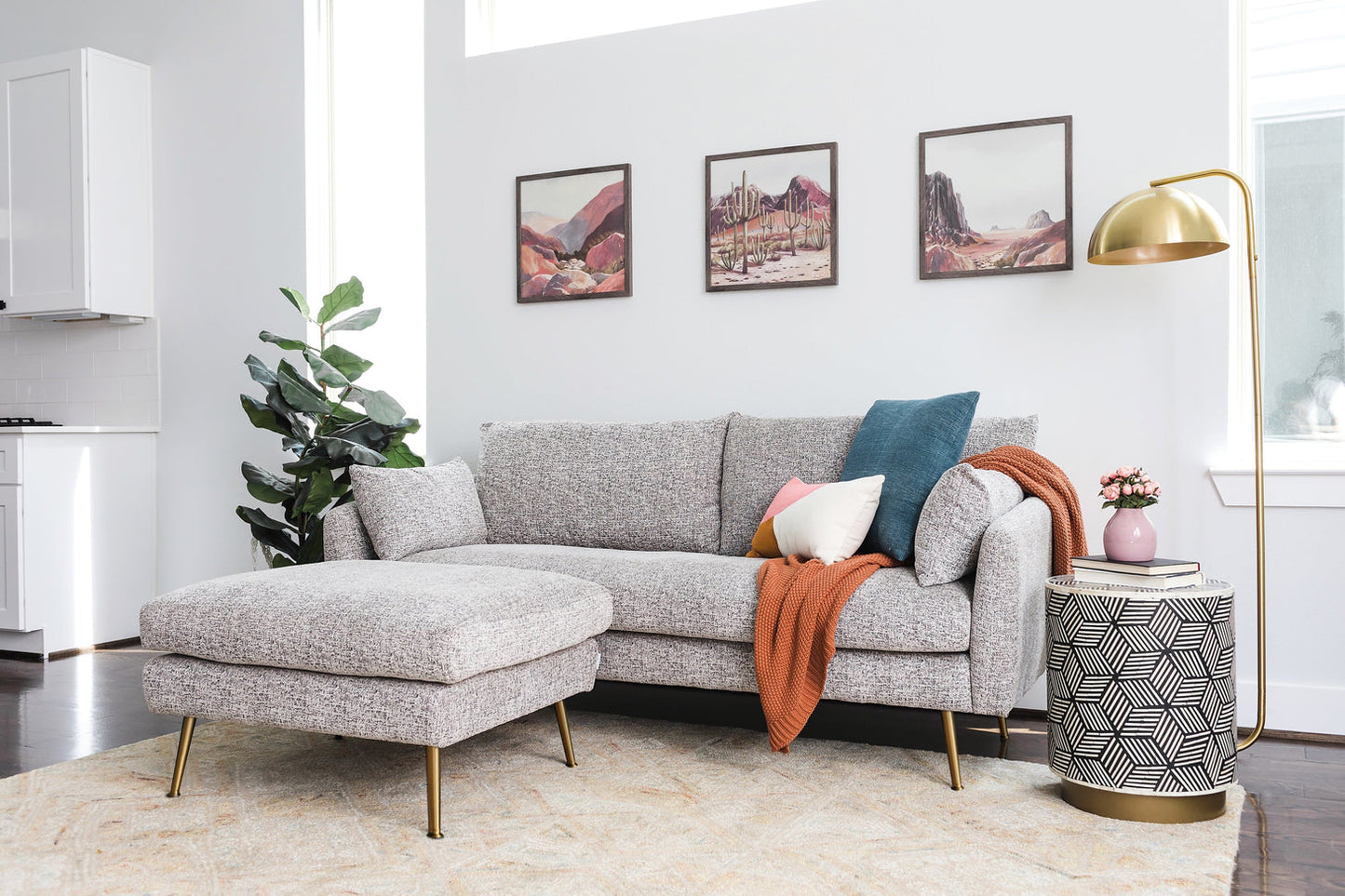 A contemporary living room featuring the Park Sofa in grey fabric, embellished with colorful pillows and an orange throw blanket. A nearby hexagonal side table holds a plant, while three landscape paintings decorate the wall. A floor lamp completes the cozy ambiance.