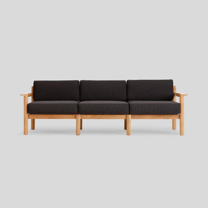 The Haven Sofa is a minimalist three-seater, featuring a light wood frame and dark cushions against a plain gray background. Its design boasts clean lines and straight armrests, capturing an elegant, modern aesthetic.