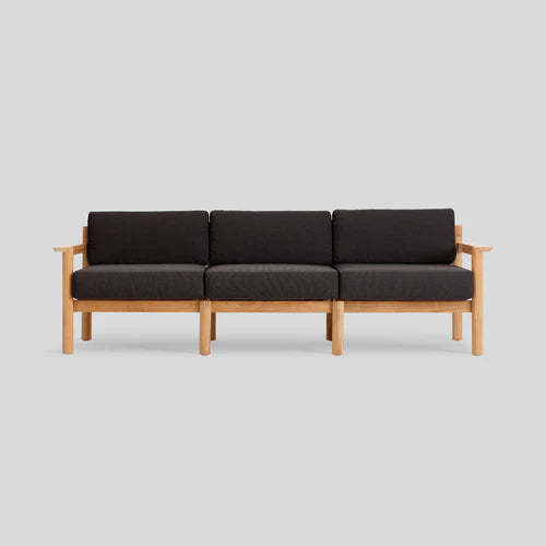 The Haven Sofa is a minimalist three-seater, featuring a light wood frame and dark cushions against a plain gray background. Its design boasts clean lines and straight armrests, capturing an elegant, modern aesthetic.