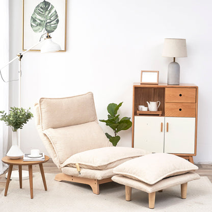 A minimalist living room features a Single Lazy Sofa Chair Leisure Foldable Reclining Chair and matching ottoman on a light rug. A wooden side table with a plant and lamp is nearby, while a wooden cabinet with a grey lamp, tea set, and framed leaf picture completes the background.