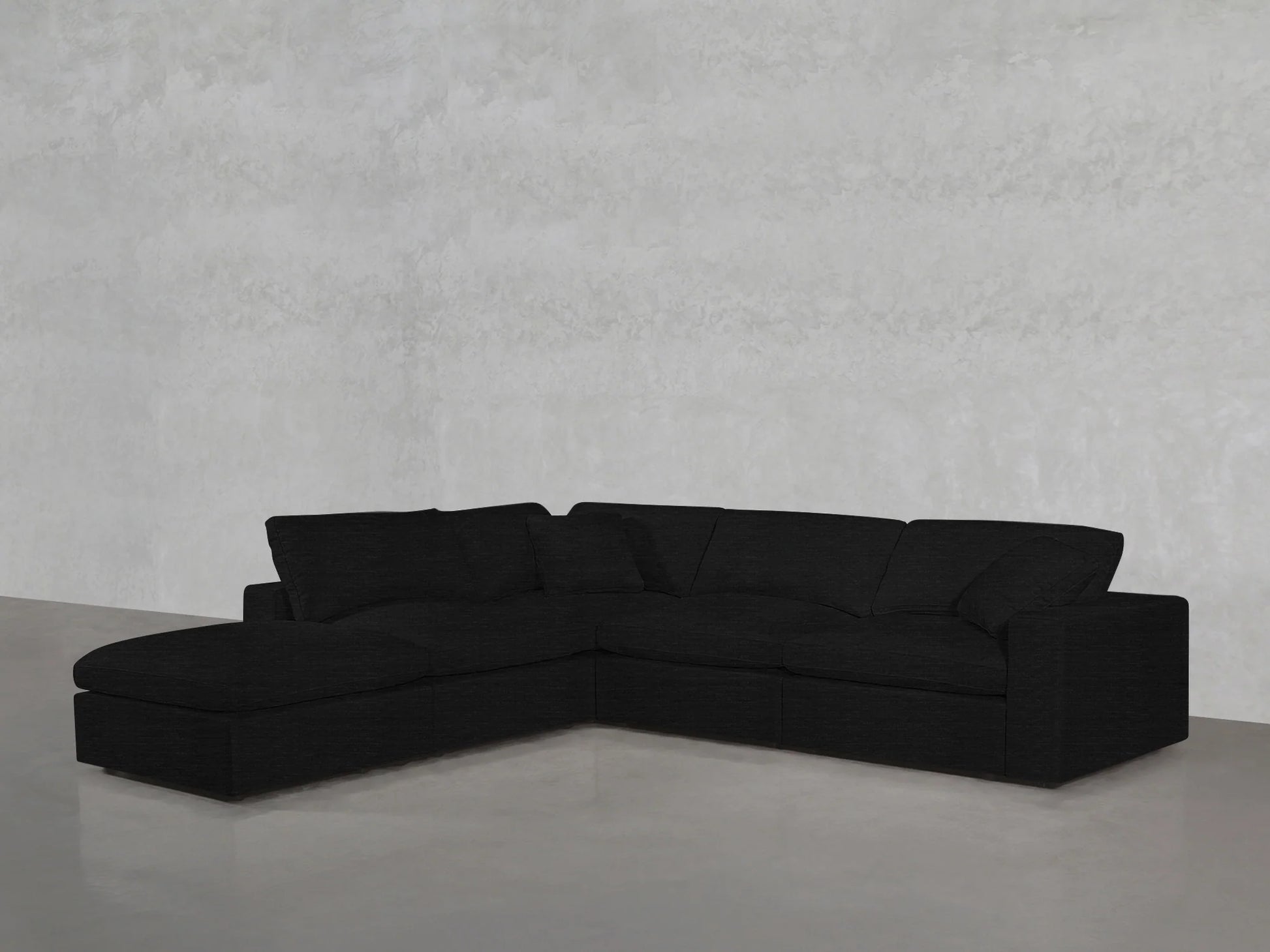 A 5-seat Modular Corner Lounger Sectional in black is placed in a minimalistic room with light gray walls and polished concrete floors. The sectional features three cushions and boasts a sleek, modern design.