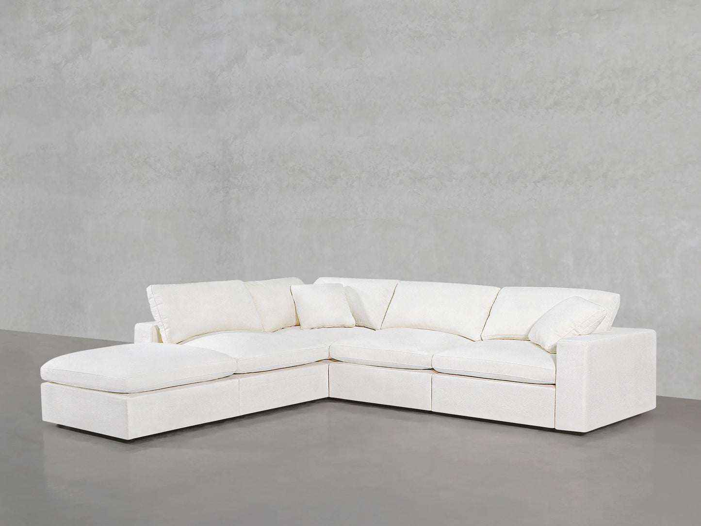 The 5-Seat Modular Corner Lounger Sectional, a spacious, contemporary off-white sofa with an L-shape design and a left-side chaise lounge extension, is placed on a sleek gray concrete floor against a simple gray concrete wall.