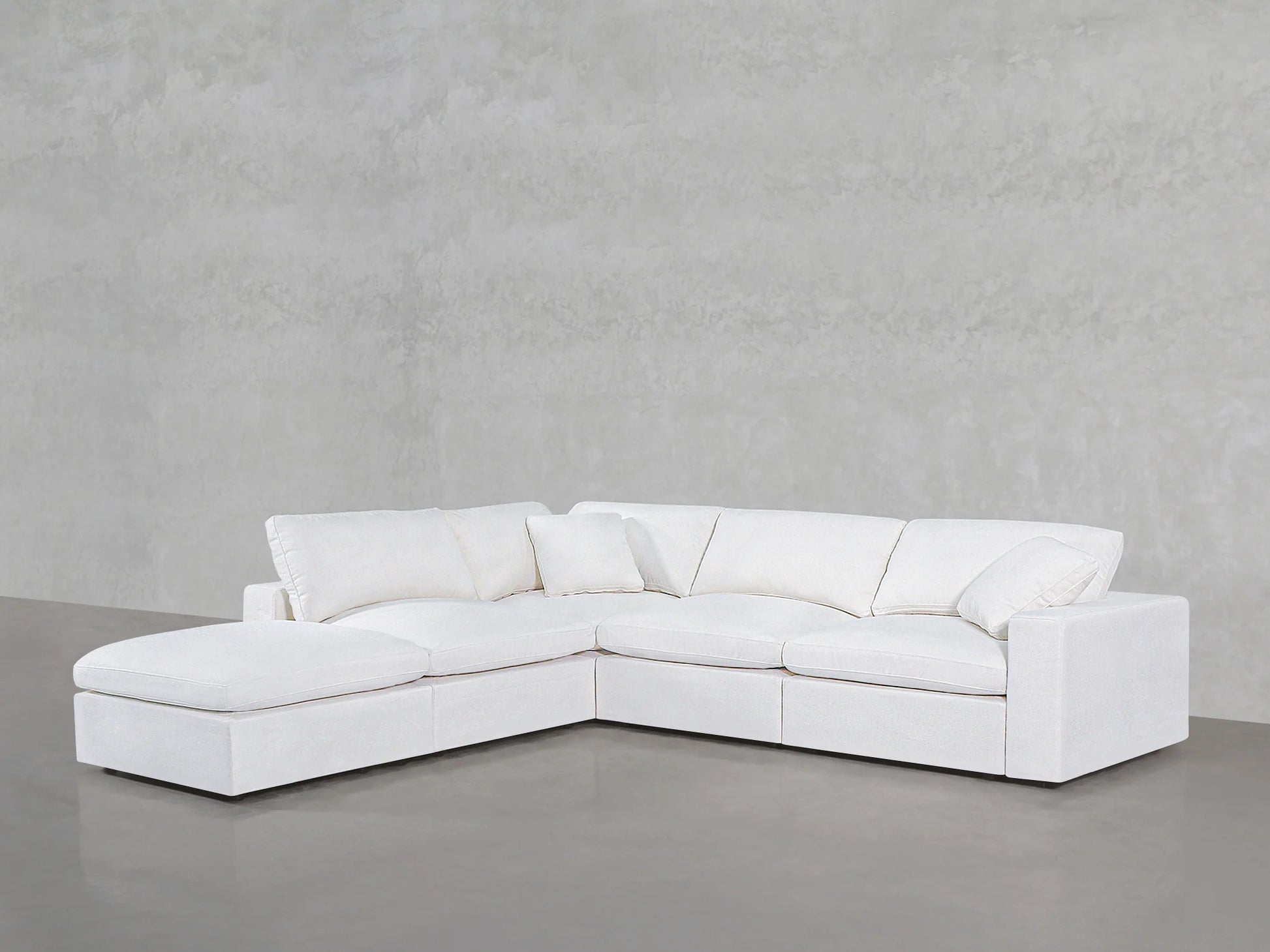 A contemporary white 5-Seat Modular Corner Lounger Sectional with plush cushions is set against a light grey concrete wall on a polished grey floor. The sectional includes a chaise on the left side, providing a spacious and comfortable seating area.