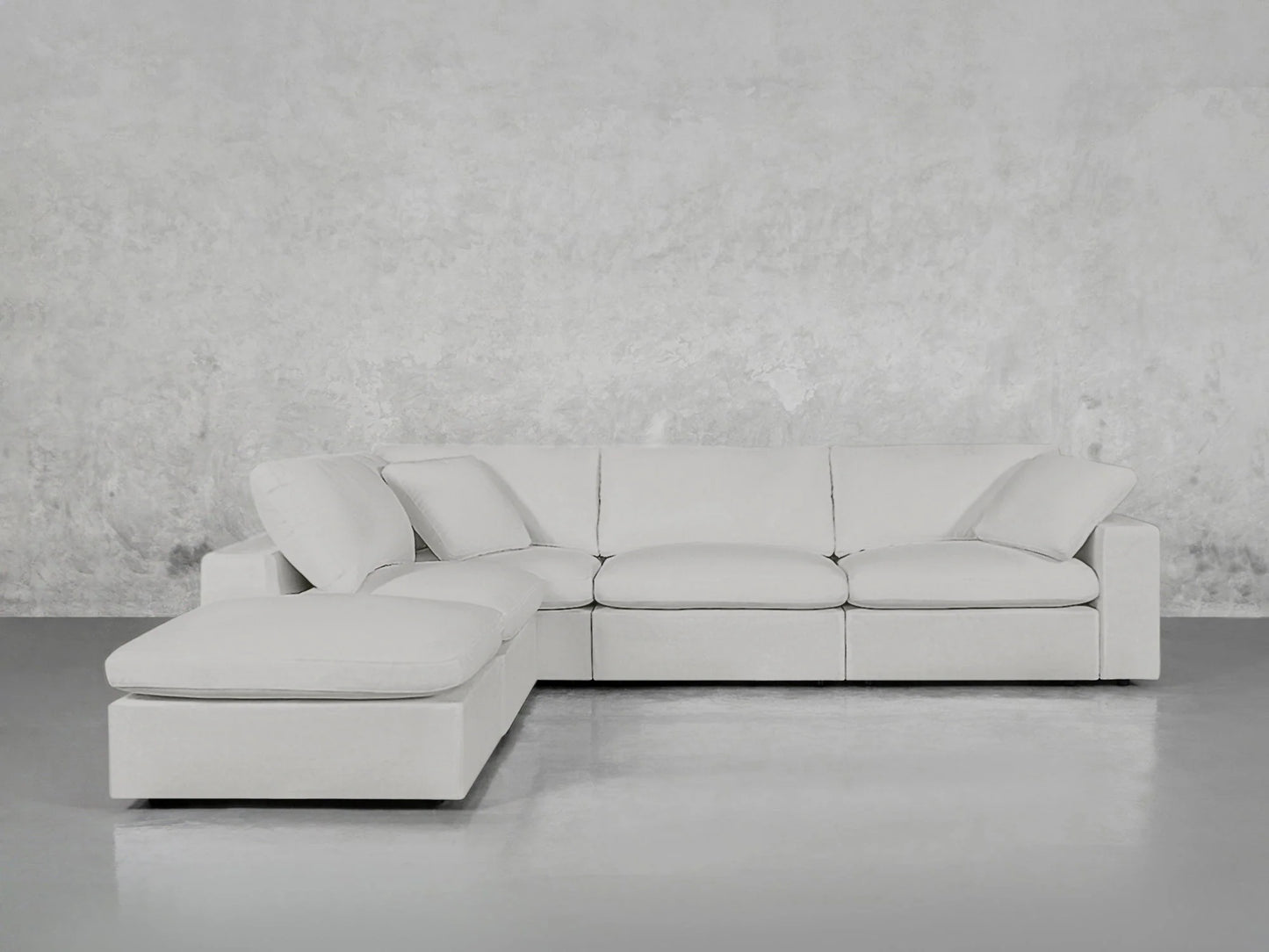 The 5-Seat Modular Corner Lounger Sectional, in light gray, is positioned against a textured gray wall on a smooth gray floor. It features plush cushions and boasts a minimalist design.