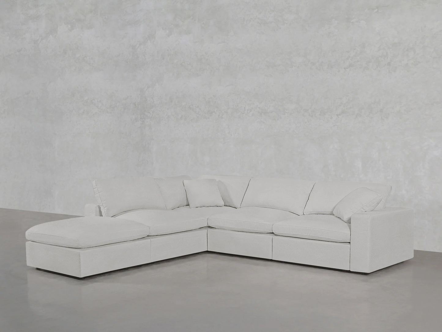 A 5-Seat Modular Corner Lounger Sectional in light gray is elegantly positioned on a smooth concrete floor, set against a textured gray wall. This modern sectional includes plush cushions and features a chaise lounge on the left side, showcasing clean lines.