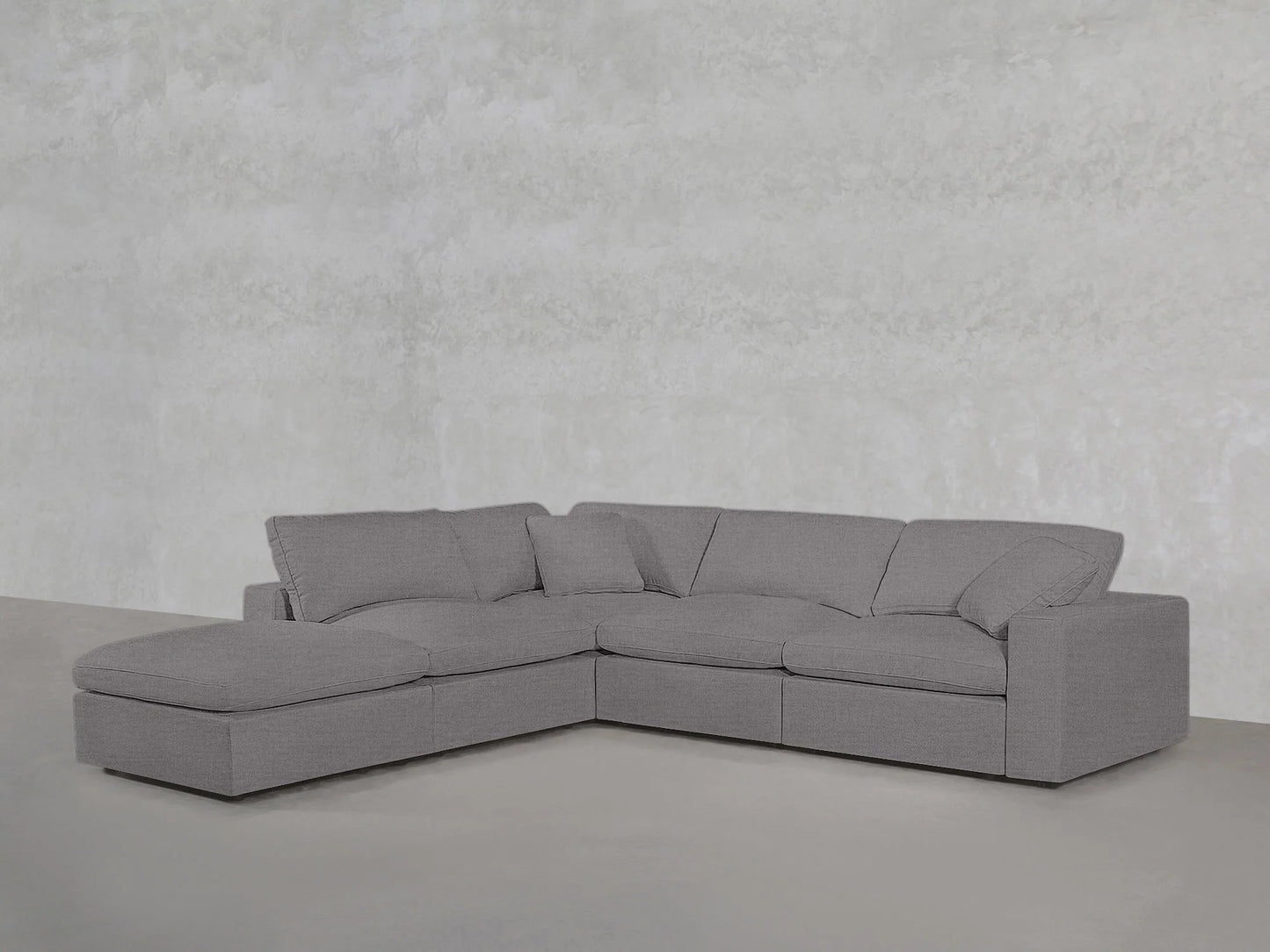 A contemporary 5-seat modular corner lounger sectional in gray, featuring a chaise lounge on the left side, set against a plain light gray wall and floor.